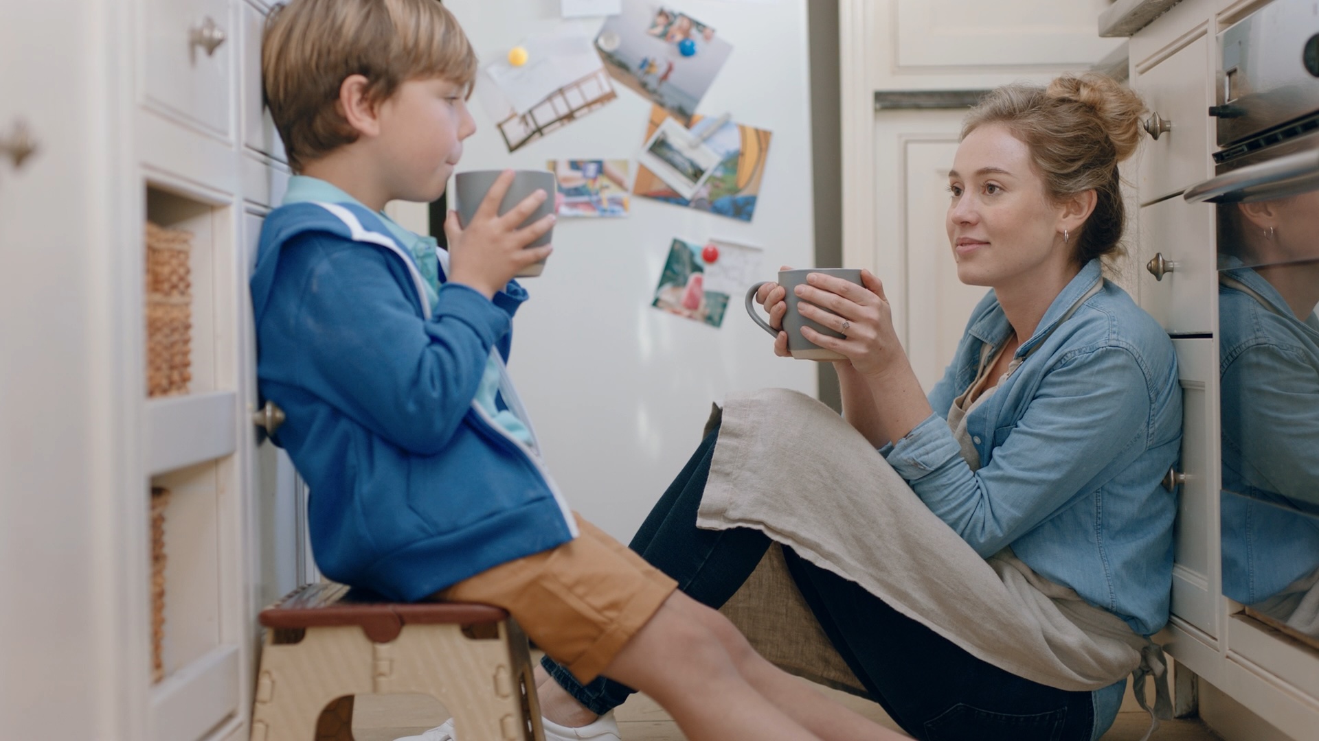 A mother and her young son sitting on the kitchen floor, talking over mugs of hot chocolate