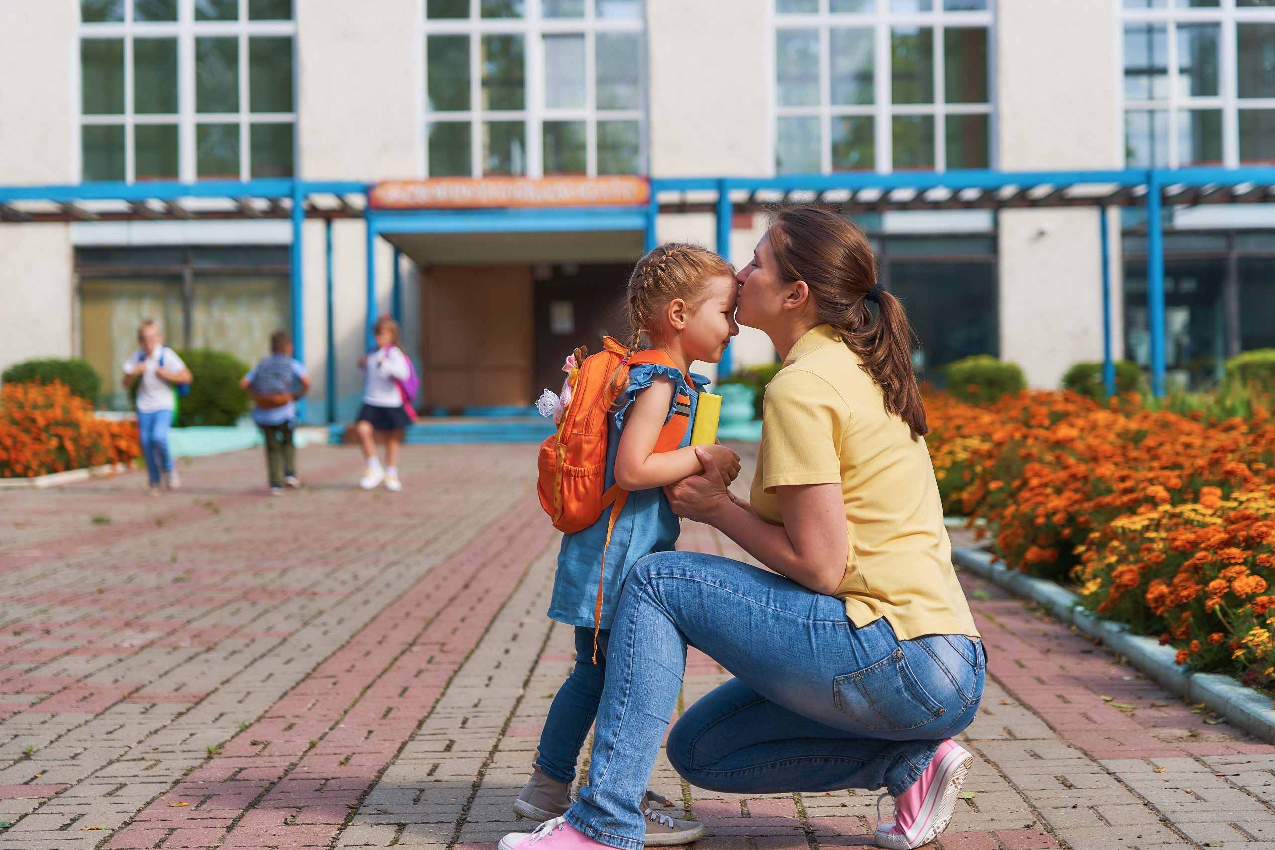 A mother kneeling to kiss her daughter goodbye outside school